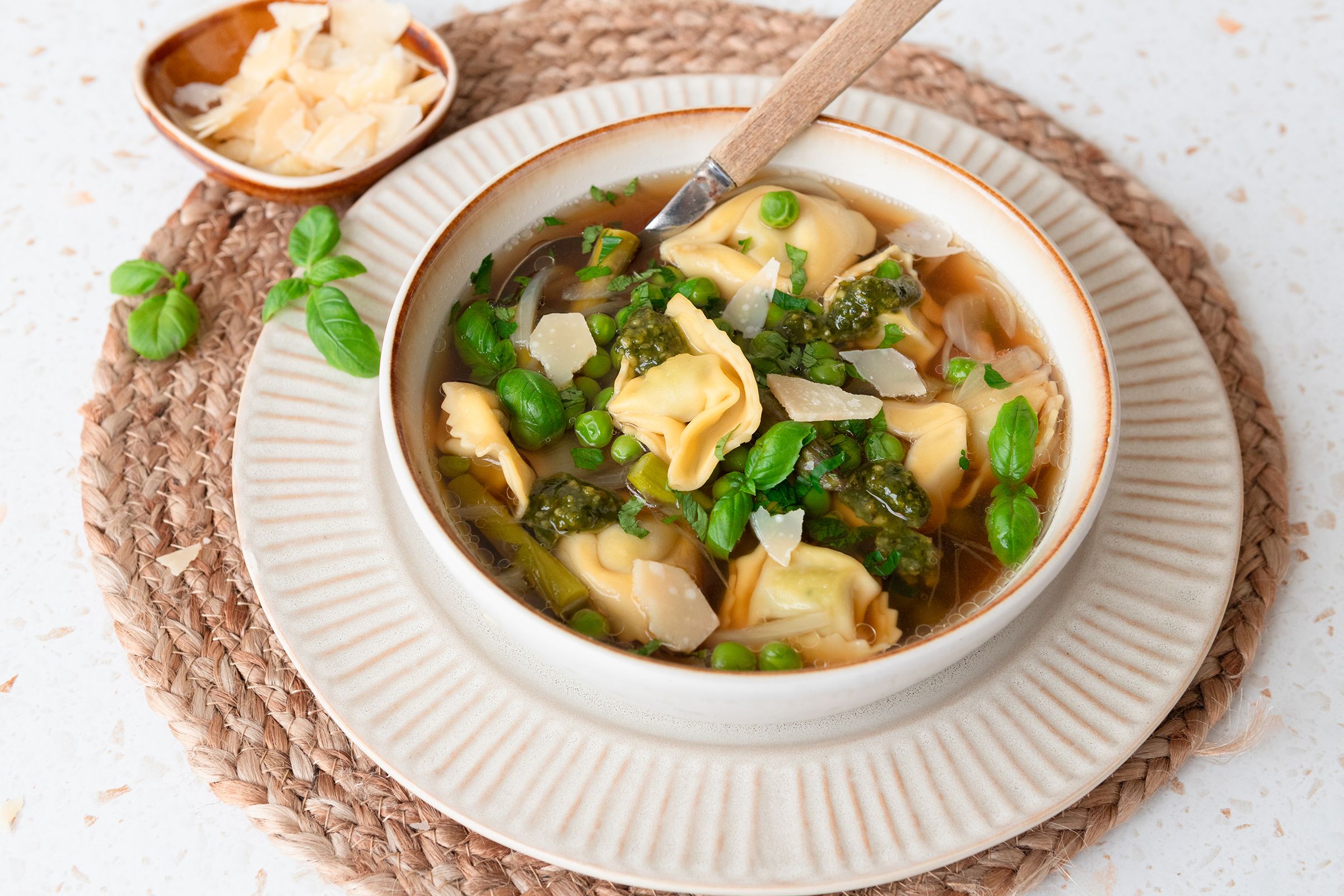 Suppe med asparges, erter og tortellini i hvit suppeskål med en skål skivet parmesan ved siden av.